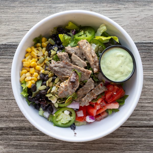 White bowl filled with steak salad, corn, black beans, tomatoes, jalapeños, and green dressing on wood.