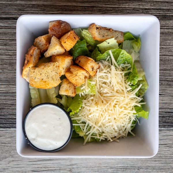 Fresh Caesar salad in a white bowl with croutons, shredded cheese, and a side of dressing.
