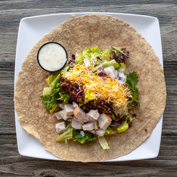 Deconstructed wrap on a large tortilla with salad, diced chicken, cheese, ground meat, and ranch.