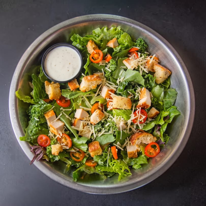 Chicken Caesar salad with croutons, shredded cheese, and cherry tomatoes in a metal bowl.