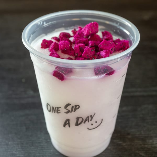 A plastic cup of light pink dragon fruit smoothie, topped with bright pink dragon fruit chunks.