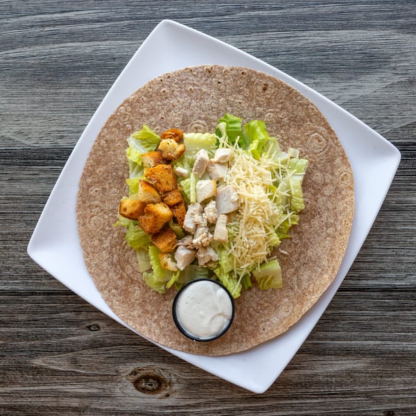 Overhead view of a chicken Caesar wrap with dressing on a white plate over a wooden table.