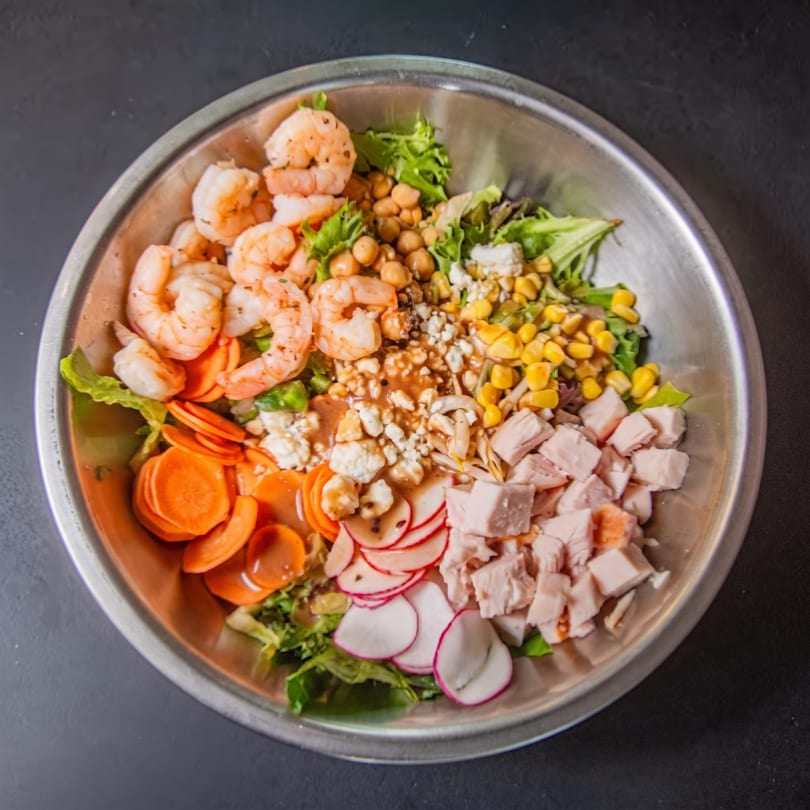 Overhead view of a vibrant salad in a metal bowl, featuring shrimp, chicken, chickpeas, corn, and radishes.