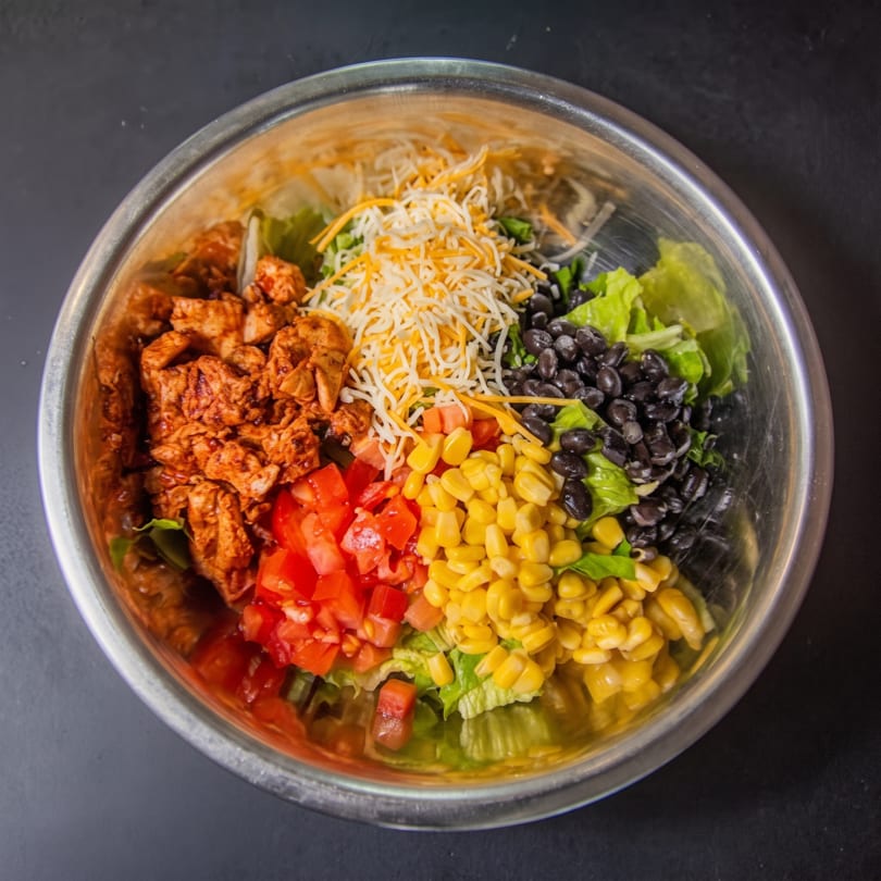 A colorful salad in a metal bowl with chicken, tomatoes, corn, black beans, cheese, and lettuce.