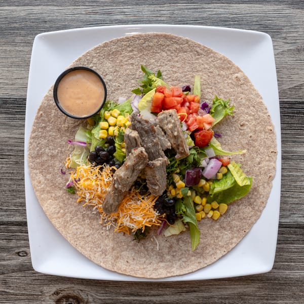 Steak wrap with lettuce, tomatoes, corn, black beans, cheese, and dressing on a white plate.