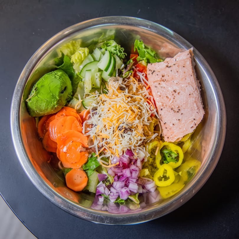 A large salad in a metal bowl with salmon, avocado, carrots, cucumbers, cheese, and onions.